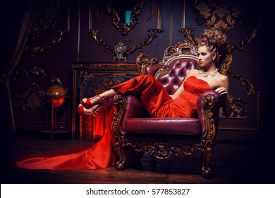 Magnificent Young Woman In Luxurious Red Dress And Precious Jewelery Is Sitting In A Chair In A Luxury Apartment. Classic Vintage Interior. Beauty, Fashion.