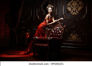 Magnificent young woman in luxurious red dress and precious jewelery posing in a luxury apartment. Classic vintage interior. Beauty, fashion.