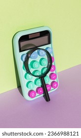 A magnifying glass and a multicolored calculator with large keys on a purple-green background. The concept of financial accounting. - Shutterstock ID 2366988121