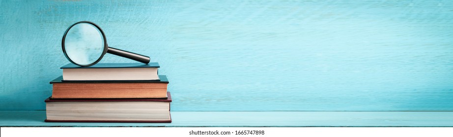 A magnifying glass on a stack of books on a blue wooden bookshelf with copy space web banner. - Shutterstock ID 1665747898