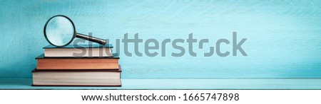 A magnifying glass on a stack of books on a blue wooden bookshelf with copy space web banner.