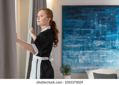 Maid Unveiling Curtains In A Hotel Room With Blue Painting On The Wall