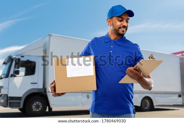 mail service and shipment concept - happy\
indian delivery man with parcel box and clipboard in blue uniform\
over truck on street\
background