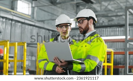 Main engineer briskly explains plan of action to employee. Bearded man holding notebook and gesturing. Workers in yellow jackets and helmets cooperate at construction site.