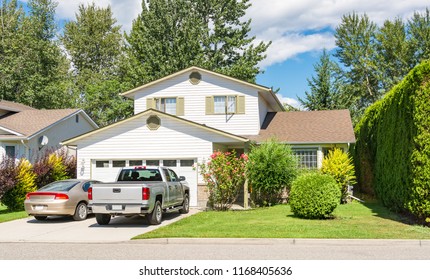 Main Entrance Of Big Residential House With Car And Truck Parked On Concrete Driveway In Front. Family House With Landscaped Front Yard On Blue Sky Background