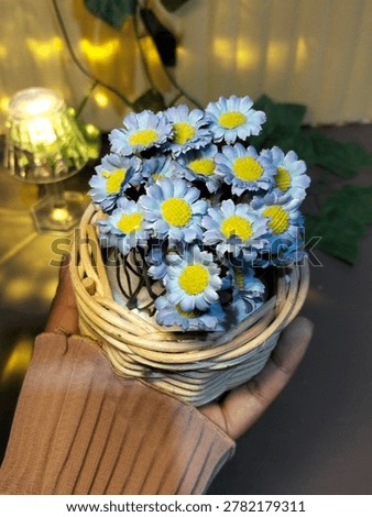 The main image shows a cluster of small, purple-blue artificial daisies with bright yellow centers (pistils). These flowers are arranged tightly in a pot.