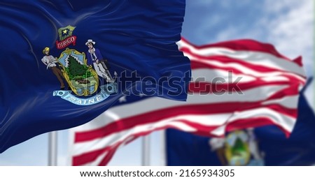 The Maine state flag waving along with the national flag of the United States of America. In the background there is a clear sky. Maine is a state in the New England region of the United States
