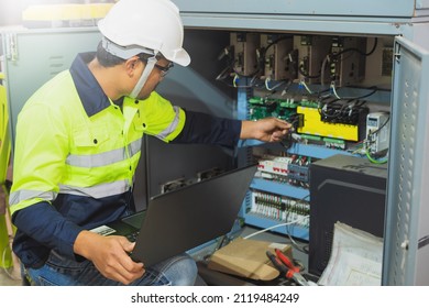 A Maintenance Engineer Wearing Green Protective Clothing Uses Laptops And Tablets To Inspect Factory Electrical Systems.