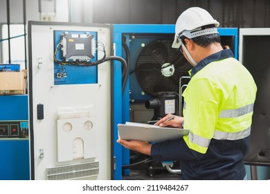 A Maintenance Engineer Wearing Green Protective Clothing Uses Laptops And Tablets To Inspect Factory Electrical Systems.