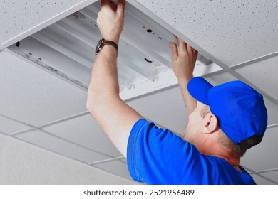 A maintenance worker in a blue cap and blue clothing is working on a ceiling-mounted fluorescent light in a commercial space, carefully adjusting the fixture as part of routine repair. - Powered by Shutterstock