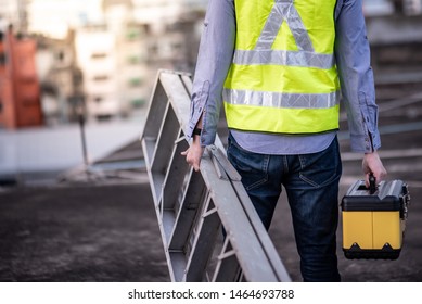 Maintenance Worker Man With Safety Helmet And Green Vest Carrying Aluminium Step Ladder And Tool Box At Construction Site. Civil Engineering, Architecture Builder And Building Service Concepts