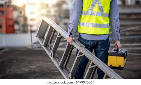 Maintenance worker man with safety helmet and green vest carrying aluminium step ladder and tool box at construction site. Civil engineering, Architecture builder and building service concepts