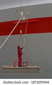 Maintenance Worker Painting Ship Hull While Hanging From Top On Platform