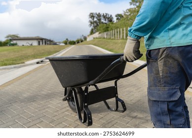 Maintenance worker pushing an empty black wheelbarrow along a paved path in a cemetery or funeral home property, performing landscaping and upkeep duties