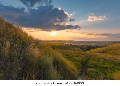 The majestic dusk scene with motley high grass and wildflowers covered hills in front of a beautiful sunset sky with clouds. Summer nature background.
