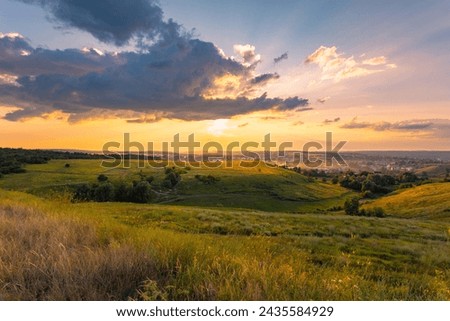 The majestic dusk scene with motley high grass and wildflowers covered hills in front of a beautiful sunset sky with clouds. Summer nature background.