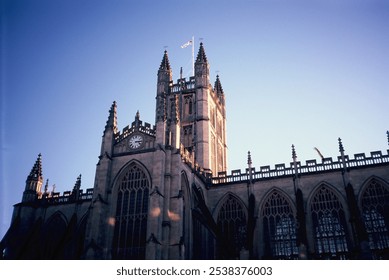 A majestic gothic-style church stands under a clear blue sky, featuring intricate arches, tall spires, and a prominent clock. The sunlight enhances the timeless elegance of the historic architecture. - Powered by Shutterstock