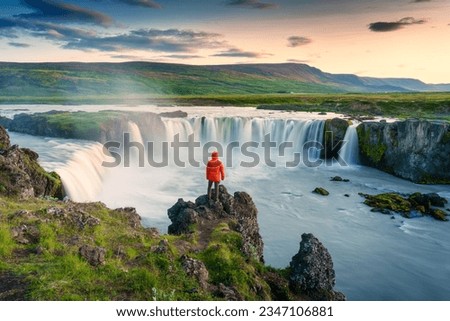 Majestic landscape of Godafoss waterfall flowing with colorful sunset sky and male tourist standing at the cliff on Skjalfandafljot river in summer at Northern Iceland