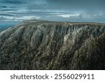 A majestic mountain, lush and verdant with trees, stands tall against a dramatic cloudy sky that looms in the background