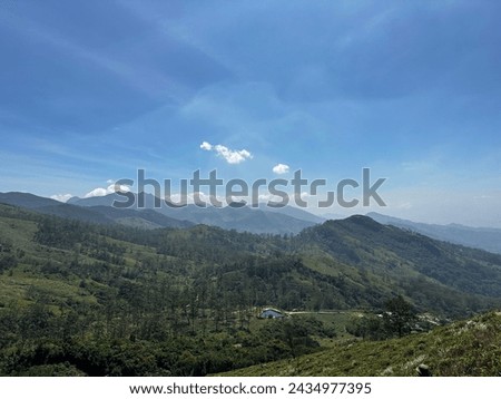 A majestic mountain range framed by lush trees, under blue skies with pure white clouds, offering a serene view with a tranquil lake—a perfect scene for hiking and embracing nature's beauty.