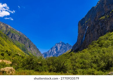 Majestic mountain range frames a deep valley with a thin waterfall cascading down a sheer cliff. Lush green vegetation and clear blue sky enhance the vibrant summer scene.