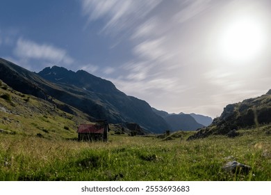 Majestic mountains with a cloudy sky, under the moonlight, with a rustic cabin on a green and flowery meadow. - Powered by Shutterstock