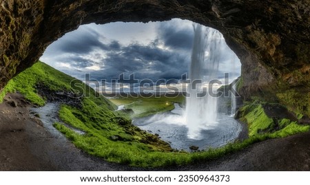Majestic natural cave with Seljalandsfoss waterfall flowing and lush field during summer on gloomy day at South of Iceland