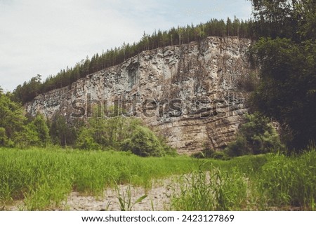 Majestic rock cliff towering over lush green forest, tranquil river, and clear blue sky. Breathtaking natural beauty, perfect for travel and nature-themed projects.