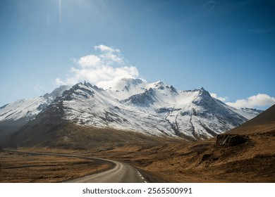 Majestic snow-capped mountain with a winding road leading into its base, framed by open plains and bright sunshine. - Powered by Shutterstock