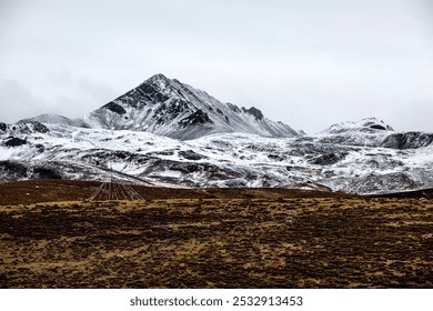 Majestic snow-dusted mountain peak under a cloudy sky, with a barren, rocky foreground featuring a lone, pyramid-like structure. - Powered by Shutterstock