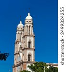 Majestic twin bell towers rise against a vibrant blue sky. Ornate architectural details adorn the white and beige facade. Historic cathedral, Mexican landmark at Culiacan, Sinaloa.