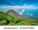 A majestic volcano in El Salvador rises above lush green hills, surrounded by clouds and a vibrant blue sky, showcasing the natural beauty and power of this dramatic landscape.