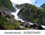 A majestic waterfall cascading down rocky cliffs surrounded by lush greenery under a clear blue sky.