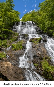a majestic waterfall over large rocks in the side of a mountain surrounded by lush green trees and plants with a gorgeous blue sky and clouds at Amicalola Falls State Park in Dawsonville Georgia USA