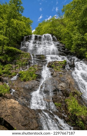 a majestic waterfall over large rocks in the side of a mountain surrounded by lush green trees and plants with a gorgeous blue sky and clouds at Amicalola Falls State Park in Dawsonville Georgia USA