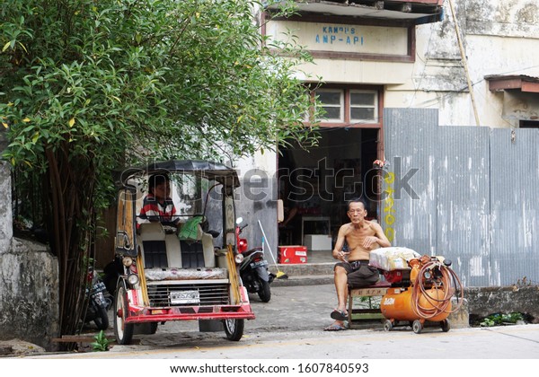MAKASSAR, INDONESIA - DECEMBER 22, 2019: An old\
Chinese man sitting in his chair while enjoying his cigarette in\
afternoon while a motorcyle rickshaw driver also park his rickshaw\
and rest.