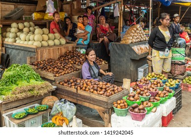 Makassar, Sulawesi, Indonesia - February 28, 2019: Terong Street Market. Female vendor sits behinds large trays of snake fruit at her booth, while a family looks on. Other veggies and fruits.