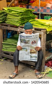 Makassar, Sulawesi, Indonesia - February 28, 2019: Terong Street Market. Older man sits at his booth reading newspaper Tribun Makassar. Folded green banana leaves in back.