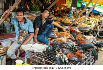 Makassar, Sulawesi, Indonesia - February 28, 2019: Terong Street Market. Two men sell living chickens, bound to iron cage, as the freshes chicken meat possible. Other food booth in back.