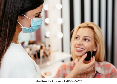 Make Up Artist With Face Protective Mask Applying Professional Make Up Of Beautiful Middle Age Blonde Woman. Coronavirus Pandemic Lifestyle.