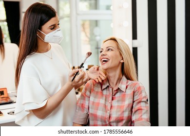 Make Up Artist With Face Protective Mask Applying Professional Make Up Of Beautiful Middle Age Blonde Woman. Coronavirus Pandemic Lifestyle.