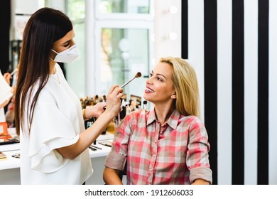 Make Up Artist With Face Protective Mask Applying Professional Make Up Of Beautiful Middle Age Blonde Woman. Coronavirus Pandemic Lifestyle.