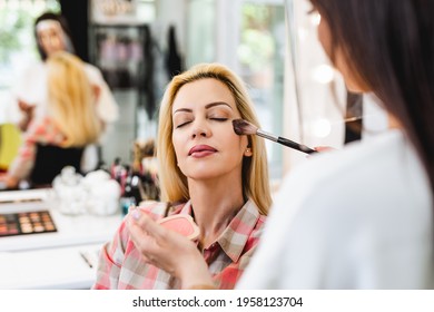 Make Up Artist With Face Protective Mask And Shield Applying Professional Make Up To Beautiful Middle Age Blonde Woman. Coronavirus Pandemic Lifestyle.