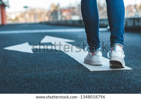 Make decision which way to go. Walking on directional sign on asphalt road. Female legs wearing jeans and white sneakers.