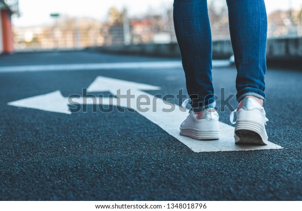 Make\
decision which way to go. Walking on directional sign on asphalt\
road. Female legs wearing jeans and white\
sneakers.