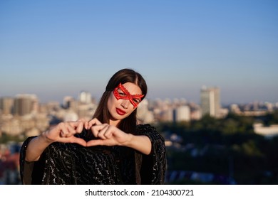 Make love, not war. Portrait of young brunette superwoman in black dress and red face mask making heart shape with hands on urban background. Freedom and equality, female power, women rights concept - Powered by Shutterstock