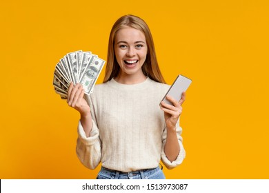 Make Money Online. Happy Teen Girl Showing Holding Smartphone And Bunch Of Money, Orange Background With Free Space