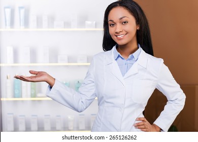 Make Your Choice. Beautiful Young African Woman In Lab Coat Keeping Arms Crossed And Smiling While Standing In Drugstore 