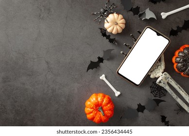 Make your Halloween greetings stand out: first person top view of skeleton hand, holding smartphone, pumpkins, eerie spiders, spiderweb, bats, bones on dark grunge backdrop, space for text or promo - Powered by Shutterstock