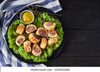 Make-ahead Party Food: Sausage Rolls Of Bulk Pork Sausage Rolled In Puff Pastry Dough On A Black Plate With Fresh Greens On A Dark Wooden Table, Top View, Copy Space